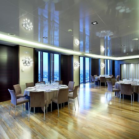 유로스타 마드리드 타워 외부 사진 The photo depicts a modern dining room or banquet hall with large windows that provide a view of the outside. The room features elegant wooden flooring and contemporary furniture, including round tables set with plates, glasses, and cutlery. Various 