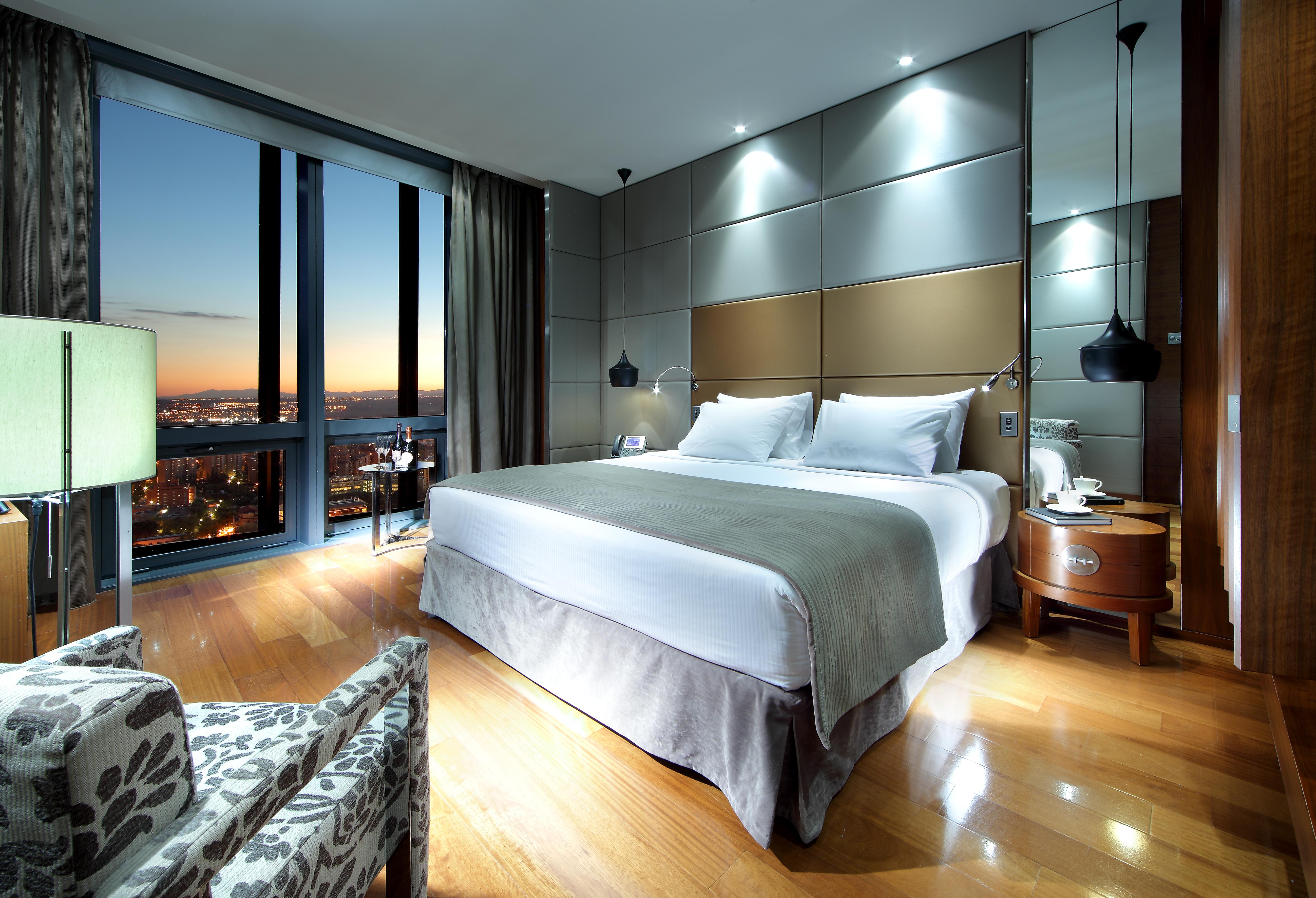 유로스타 마드리드 타워 외부 사진 The image shows a modern hotel room featuring a large bed with white linens and a gray throw. There are stylish bedside lamps and a sleek nightstand. A comfortable chair with a patterned fabric sits to the side. The room has large windows that reveal