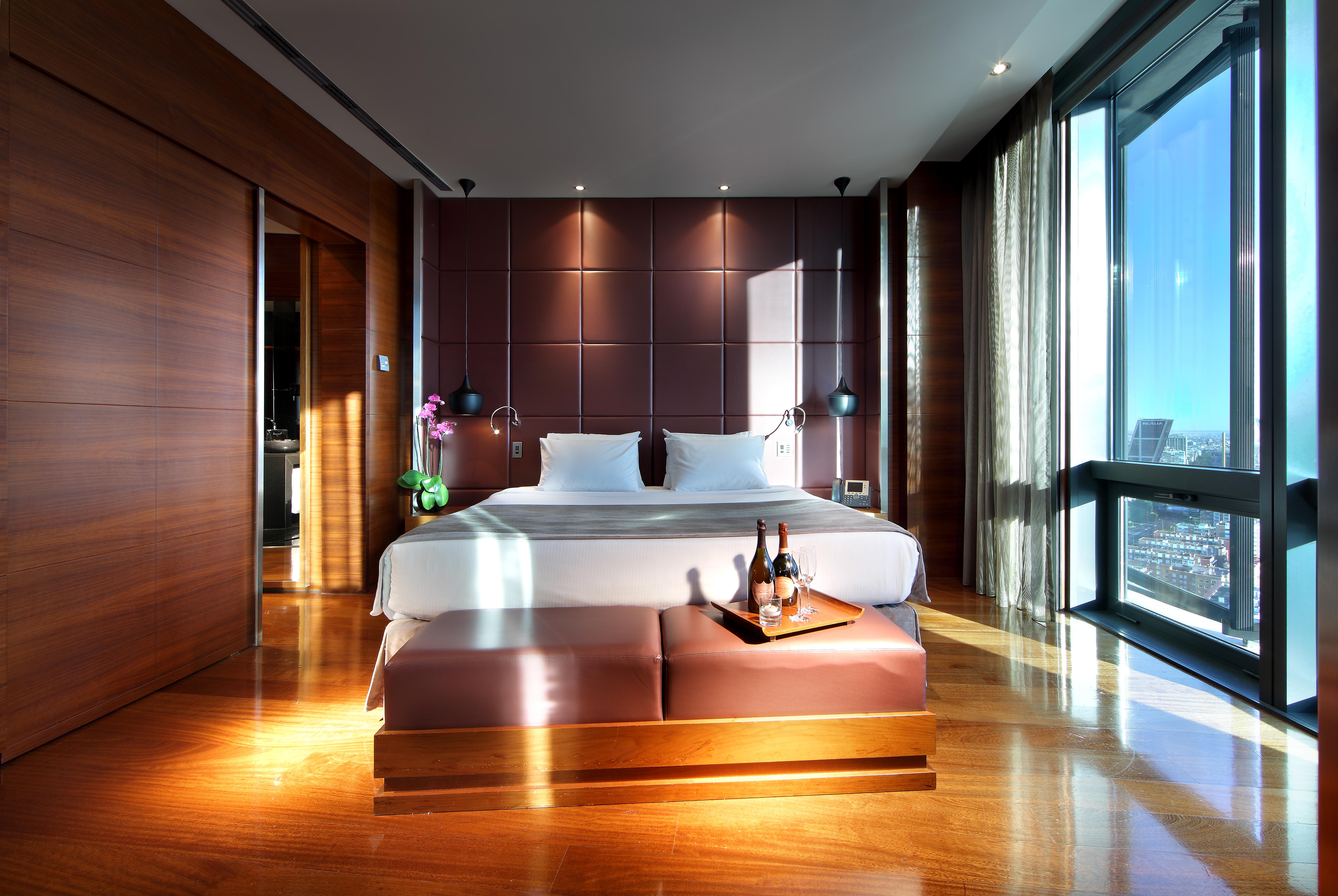 유로스타 마드리드 타워 외부 사진 The photo shows a modern hotel room featuring a large bed with white linens positioned against a stylish dark brown accent wall. There are two bedside lamps on either side of the bed, and a small table at the foot of the bed holding a bottle of wine 