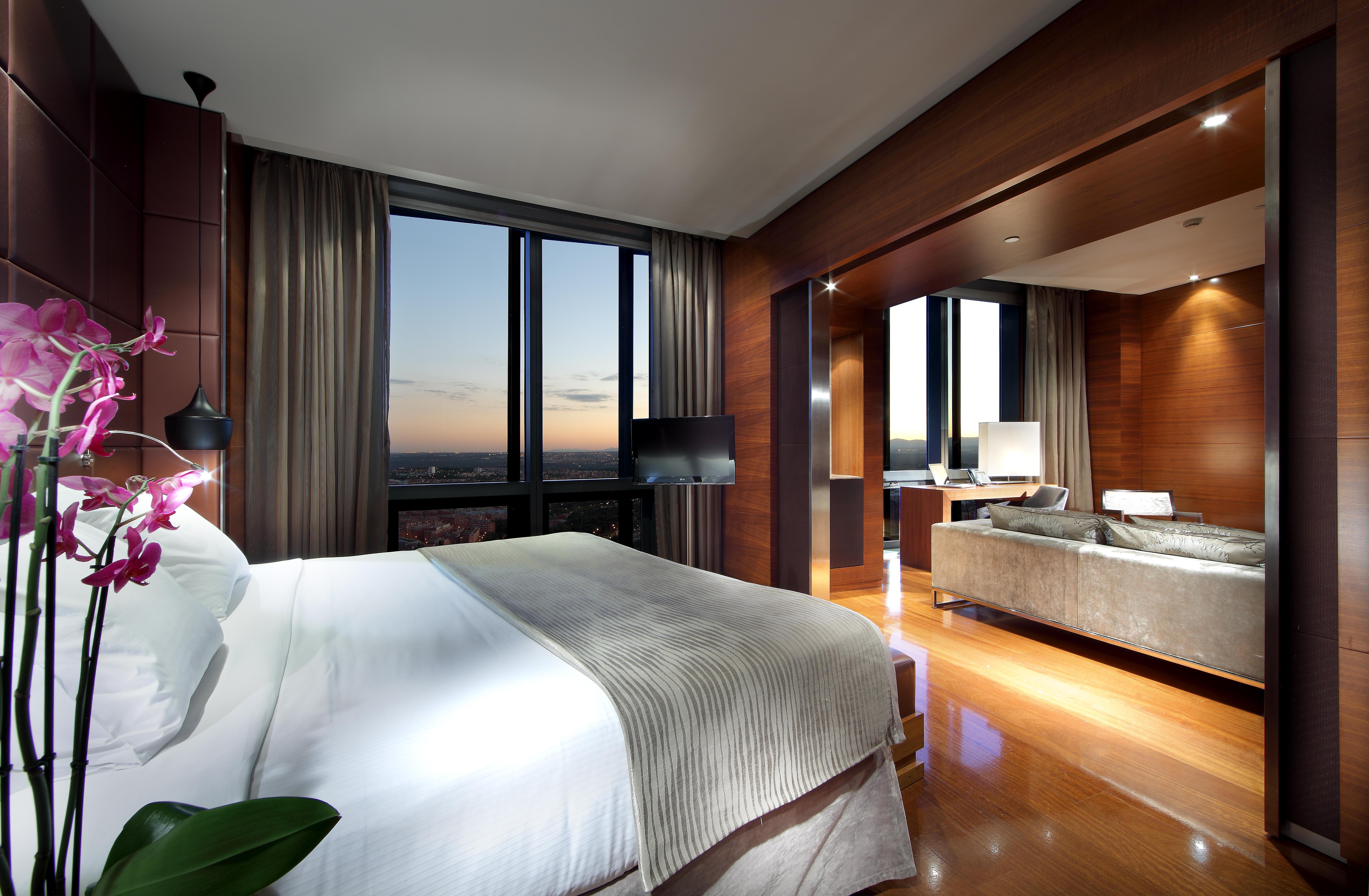 유로스타 마드리드 타워 외부 사진 The photo depicts a modern hotel room interior. It features a spacious layout with a large bed dressed in white linens, accompanied by a subtle patterned blanket. To the side, there is a small decorative plant. 

The room is illuminated by ambient li