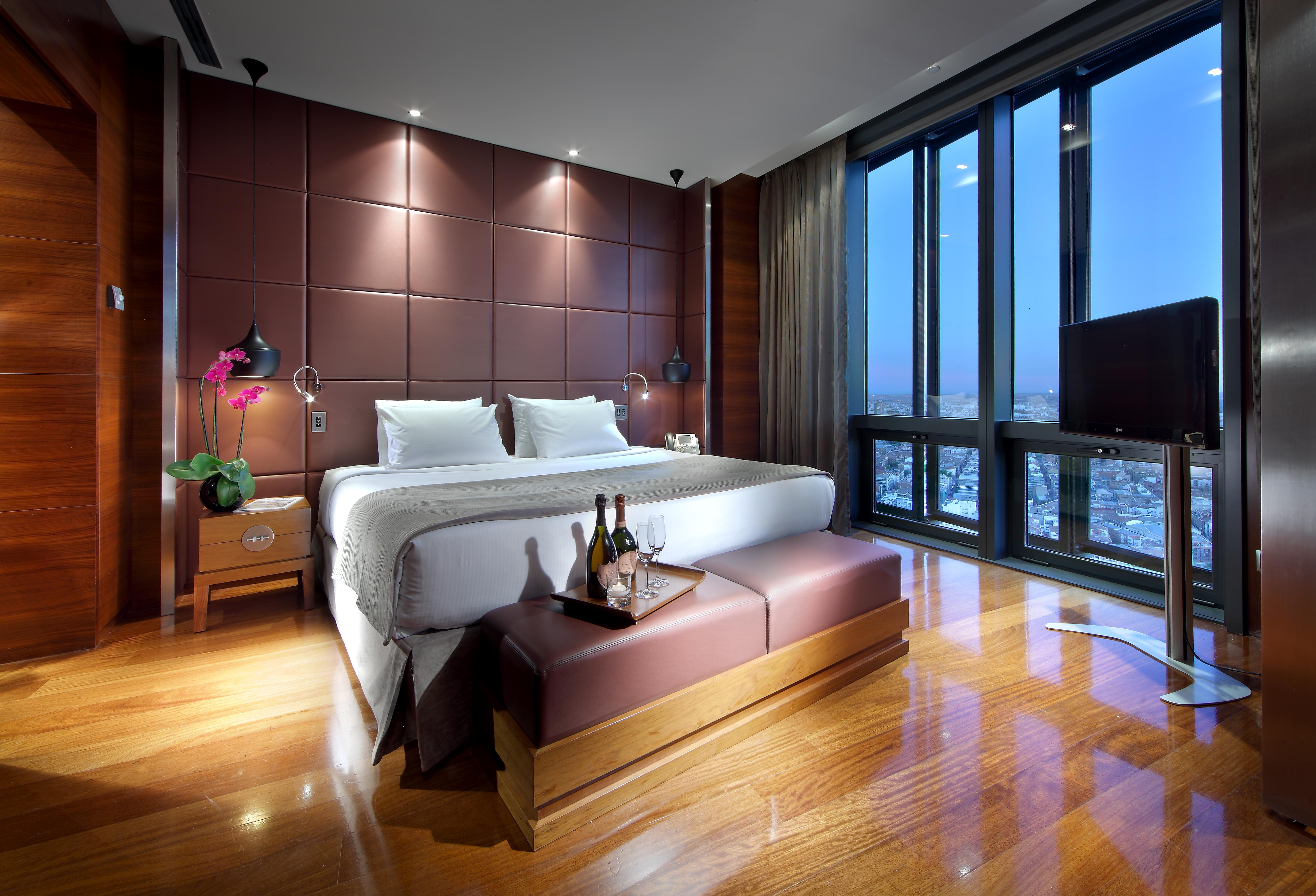 유로스타 마드리드 타워 외부 사진 The photo shows a modern hotel room with a luxurious design. It features a large bed with white linens and plush pillows, positioned against a stylish brown accent wall. There are bedside tables on each side of the bed, each equipped with a lamp. In 