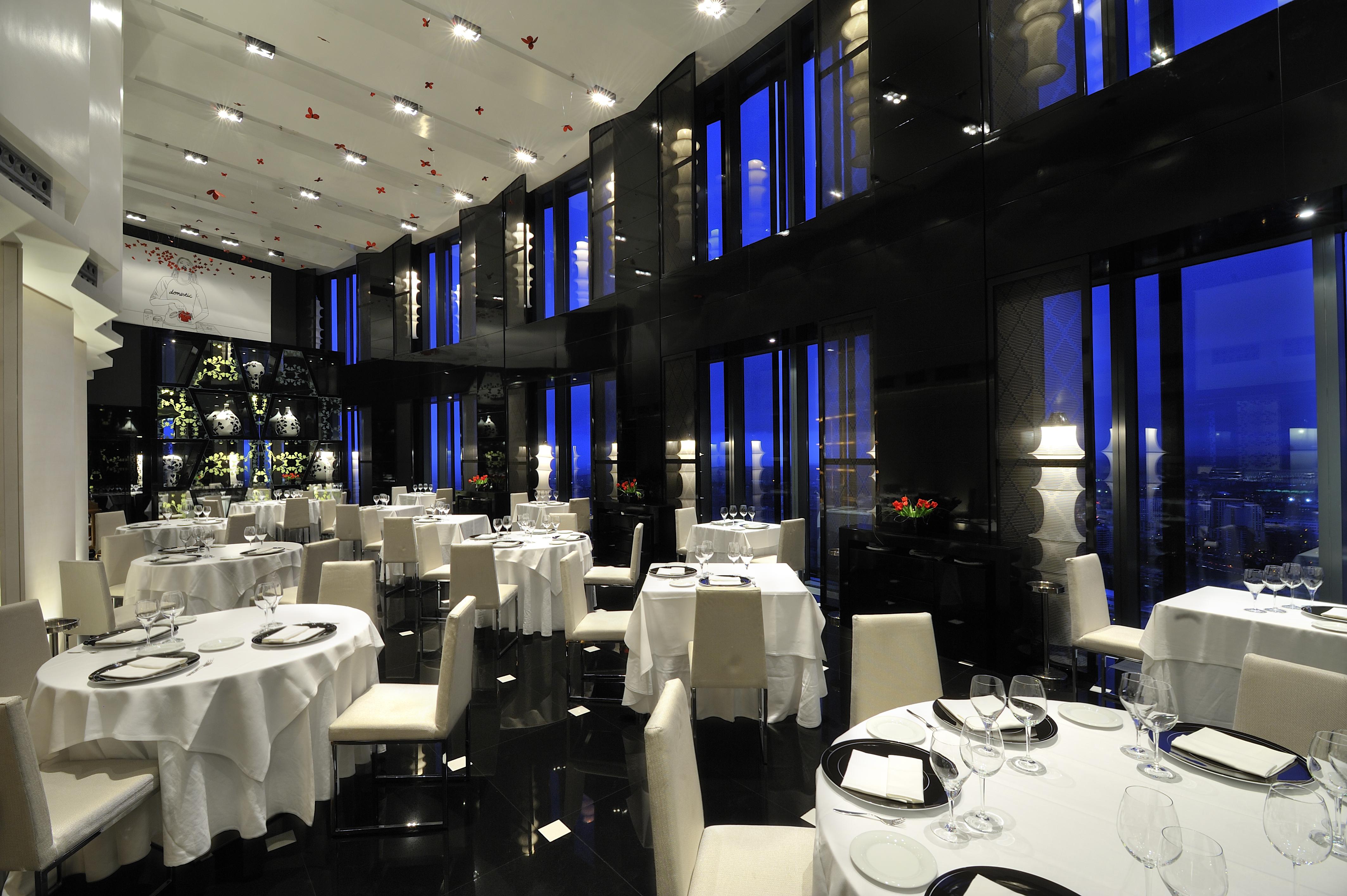 유로스타 마드리드 타워 외부 사진 The photo shows an elegant restaurant interior, characterized by its modern design and luxurious ambiance. The space features several round dining tables, each set with white tablecloths and tableware, including wine glasses. The walls are made of da