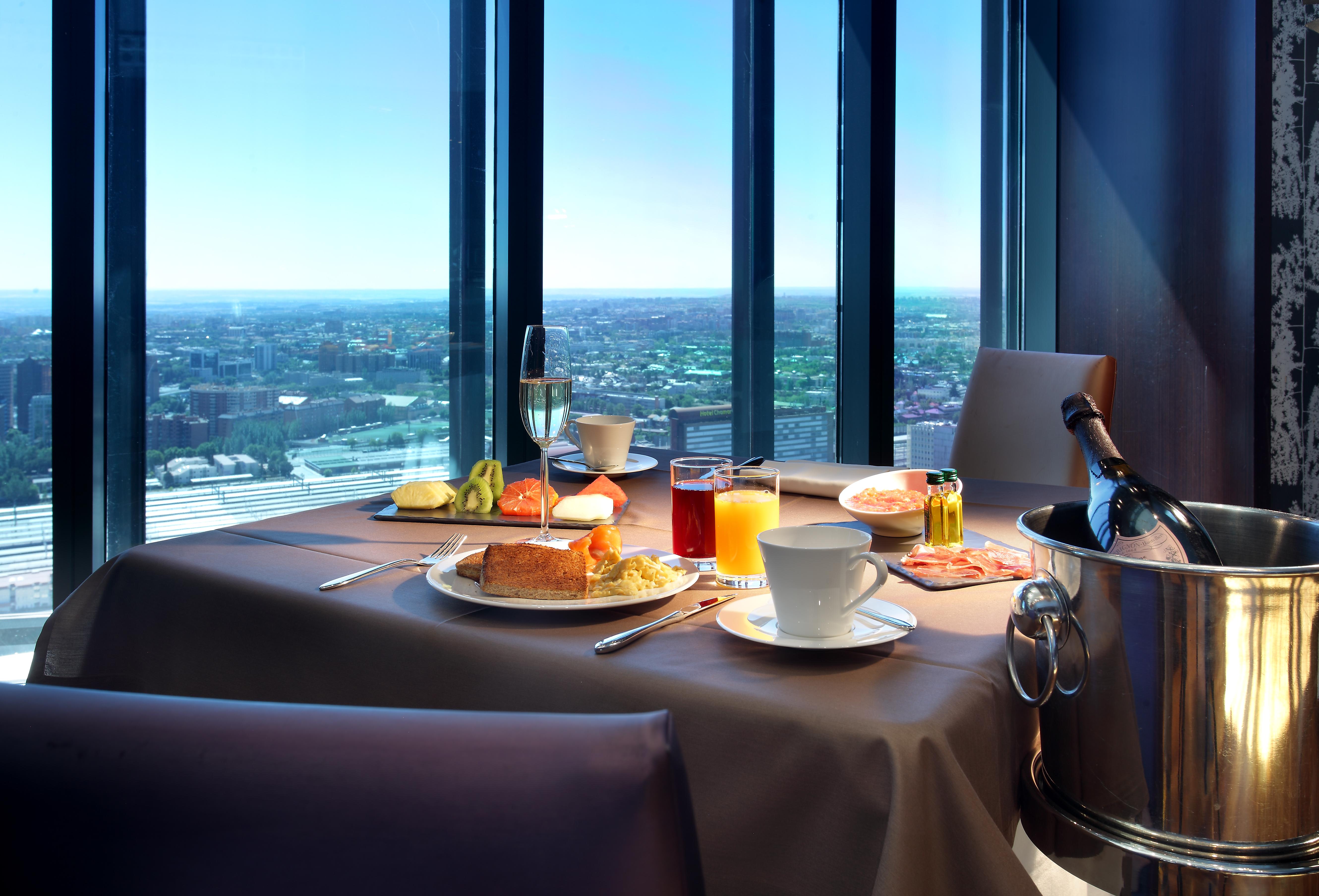 유로스타 마드리드 타워 외부 사진 The photo shows a beautifully set table with a scenic view through large windows. On the table, there are various food items, including a breakfast plate with items like eggs, pastries, and fruit. There are drinks, including a cup of coffee, a glass 