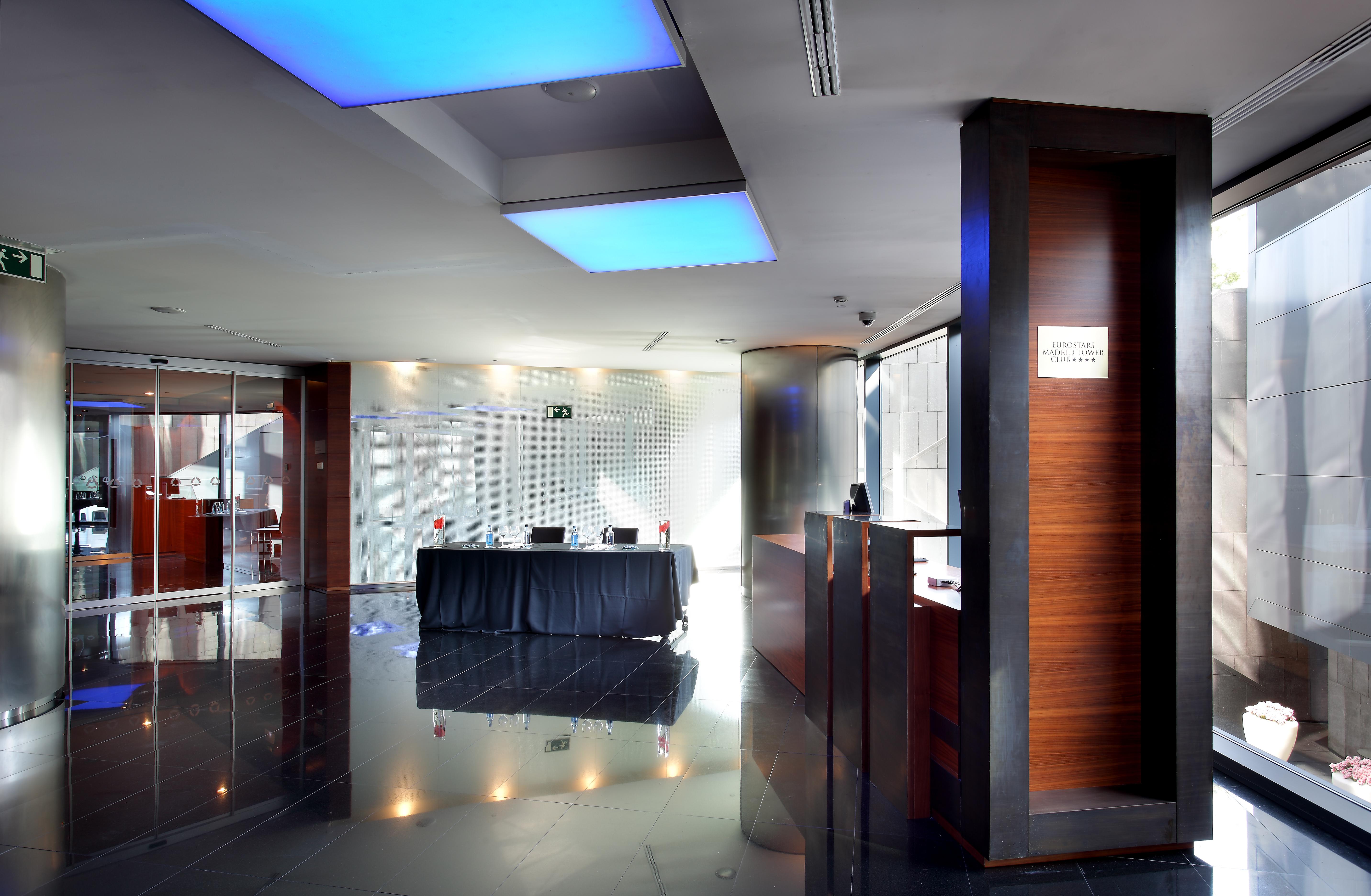 유로스타 마드리드 타워 외부 사진 The image shows the interior of a modern reception area. The space features sleek design elements, including glossy black flooring and walls. There is a front desk made of wood, which includes a sign, likely indicating reception services. Soft blue l
