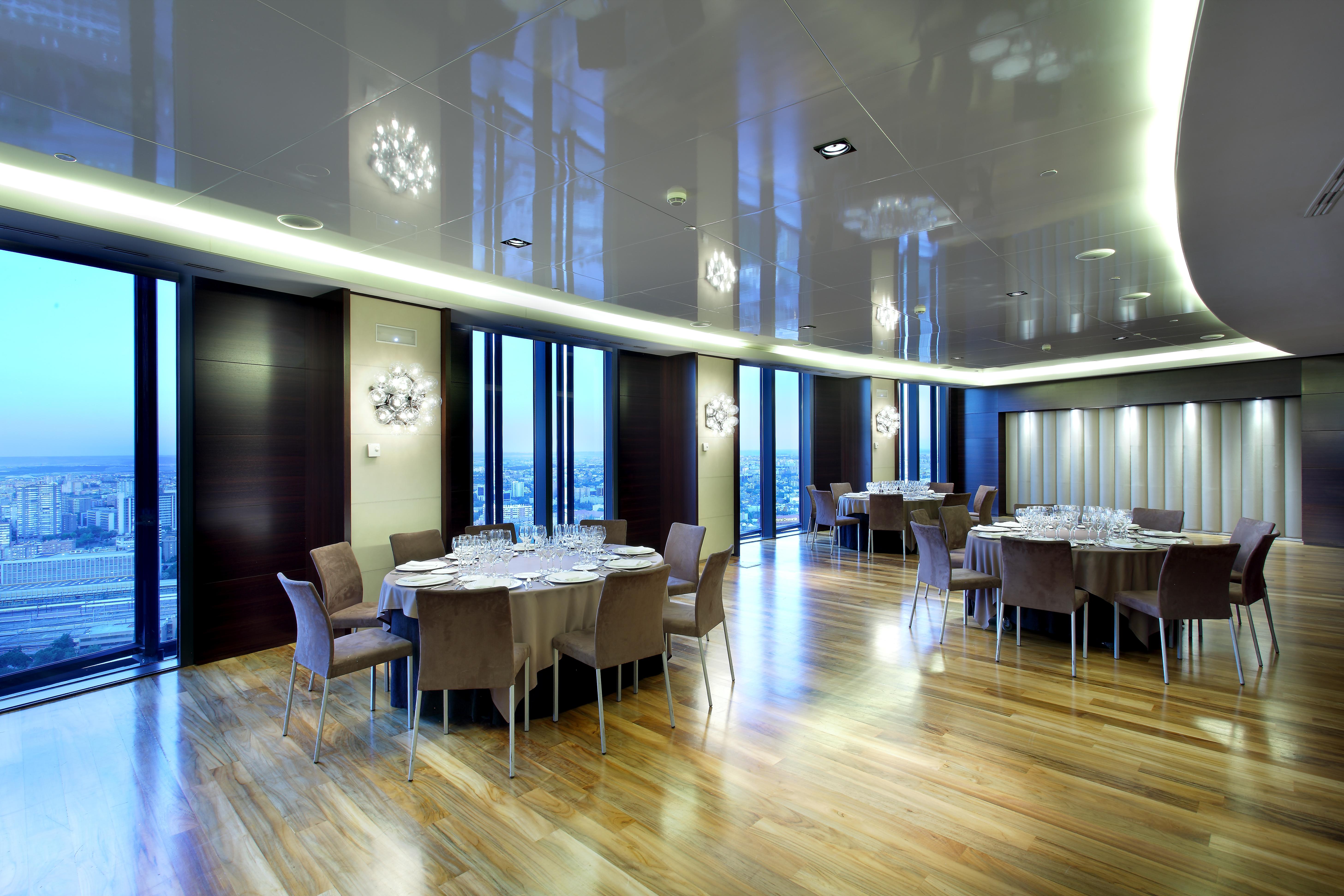 유로스타 마드리드 타워 외부 사진 The photo depicts a modern dining room or banquet hall with large windows that provide a view of the outside. The room features elegant wooden flooring and contemporary furniture, including round tables set with plates, glasses, and cutlery. Various 