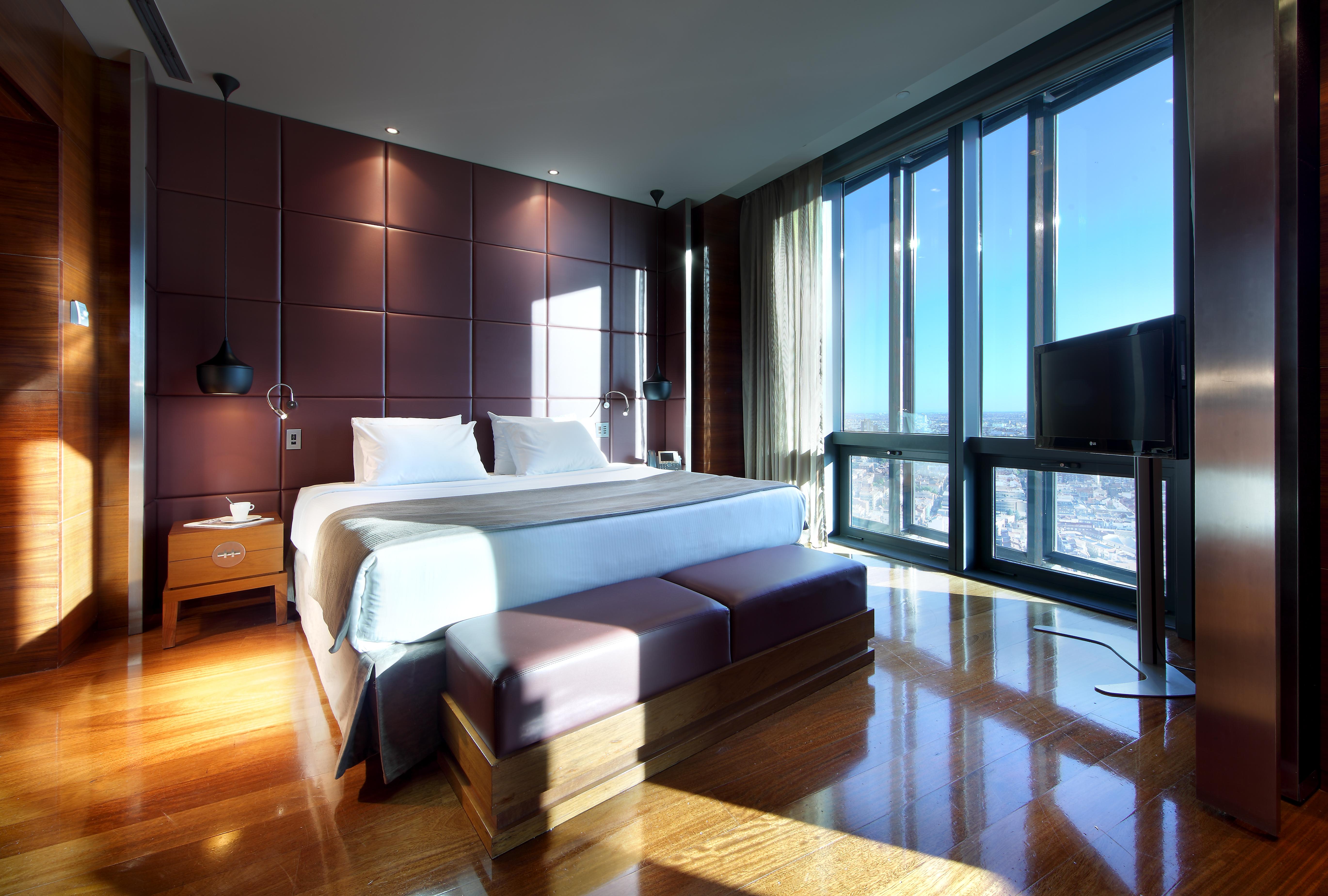 유로스타 마드리드 타워 외부 사진 The photo shows a modern hotel room with sleek design features. It includes a large bed with white bedding, positioned against a wall with dark, textured panels. On either side of the bed, there are bedside tables with lamps. A stylish bench sits at 