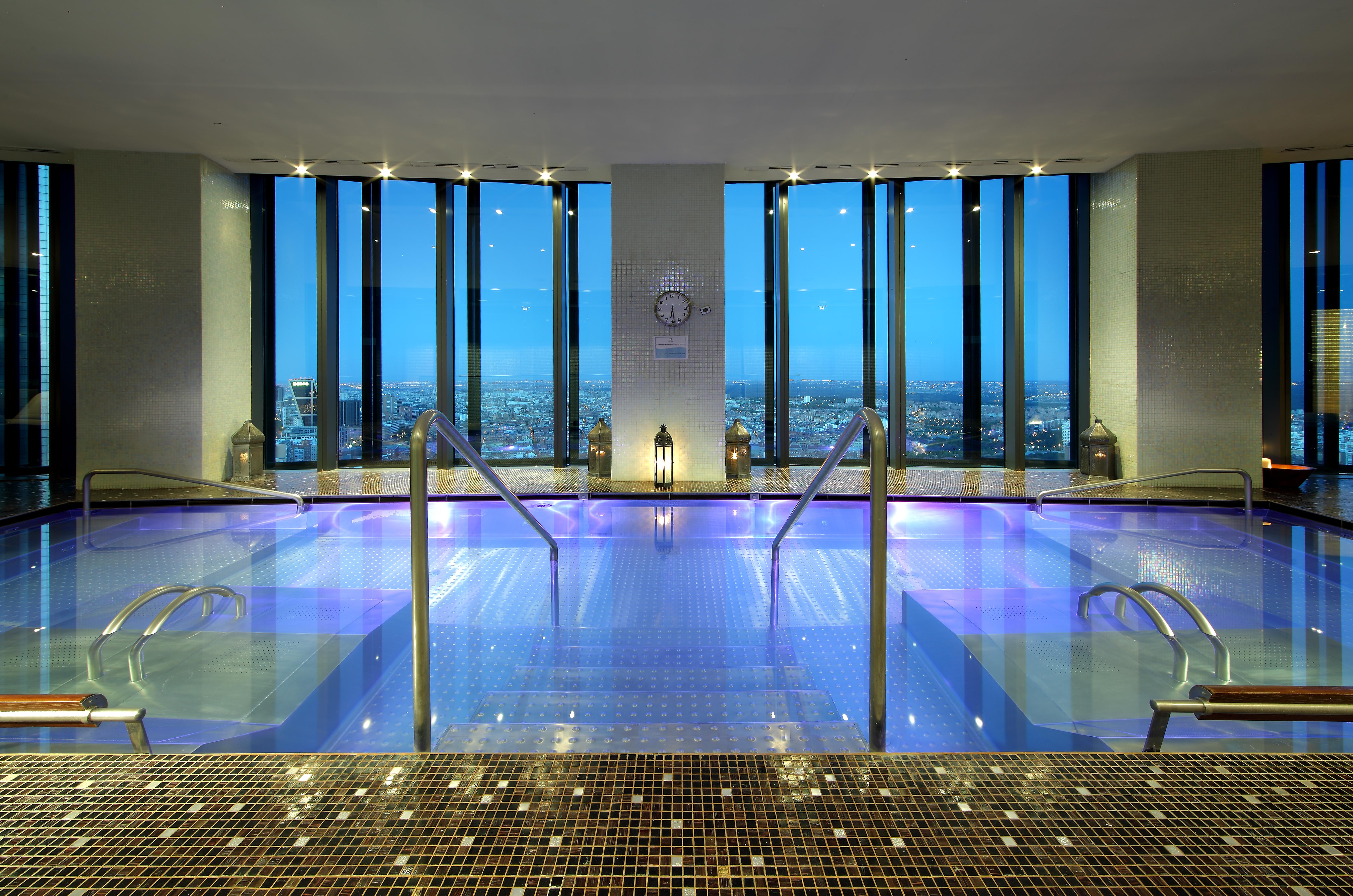 유로스타 마드리드 타워 외부 사진 The image shows a luxurious spa area with a large, tranquil pool that has a vibrant blue hue. The pool is surrounded by sleek, modern design elements, including steps leading down into the water. Large windows provide a stunning view of the cityscape