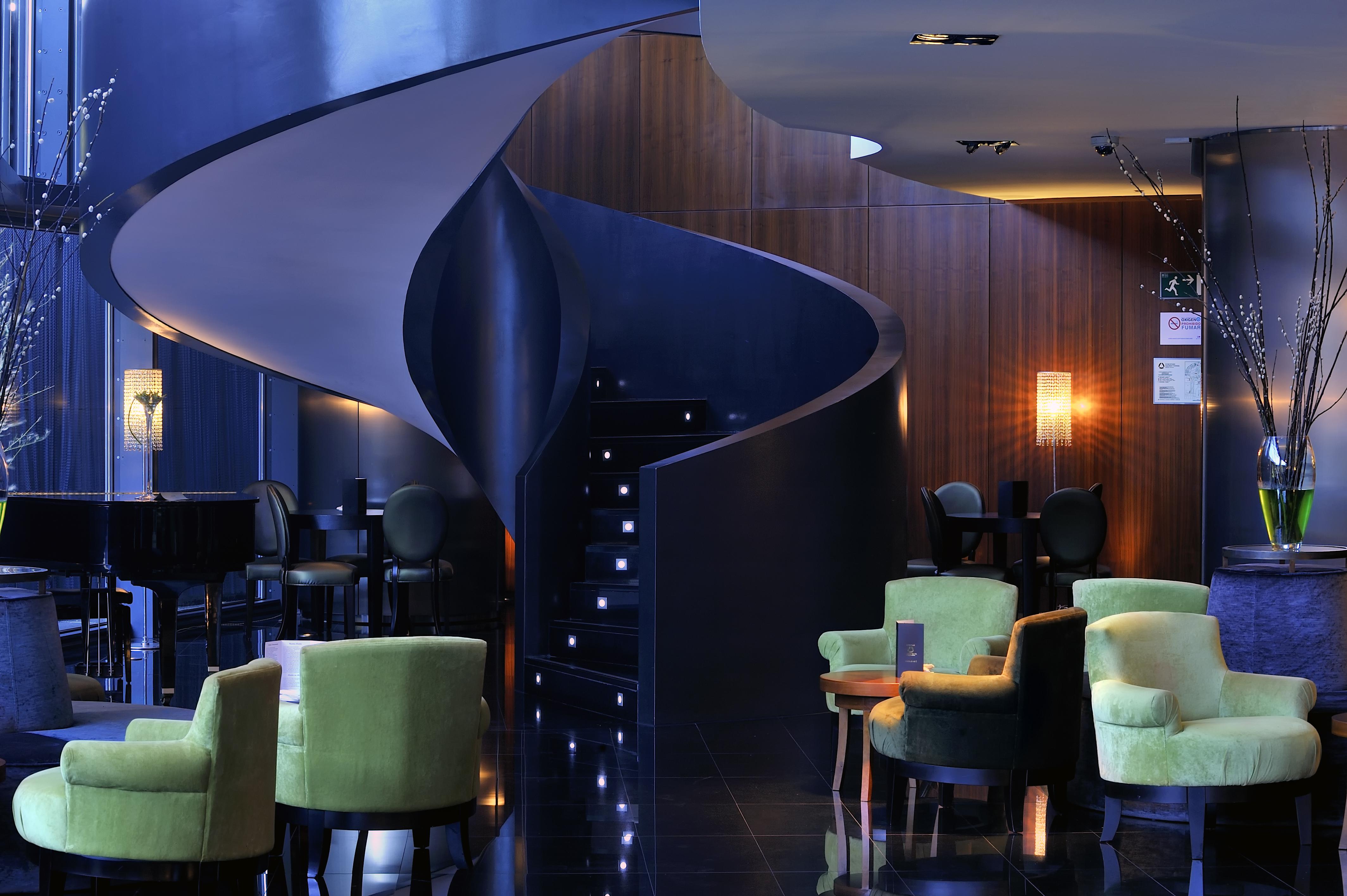 유로스타 마드리드 타워 외부 사진 The photo shows a stylish interior of a modern lounge or hotel lobby. There is a striking spiral staircase that leads to an upper level, illuminated by soft lights along the steps. The color palette features dark tones, including a deep blue wall and