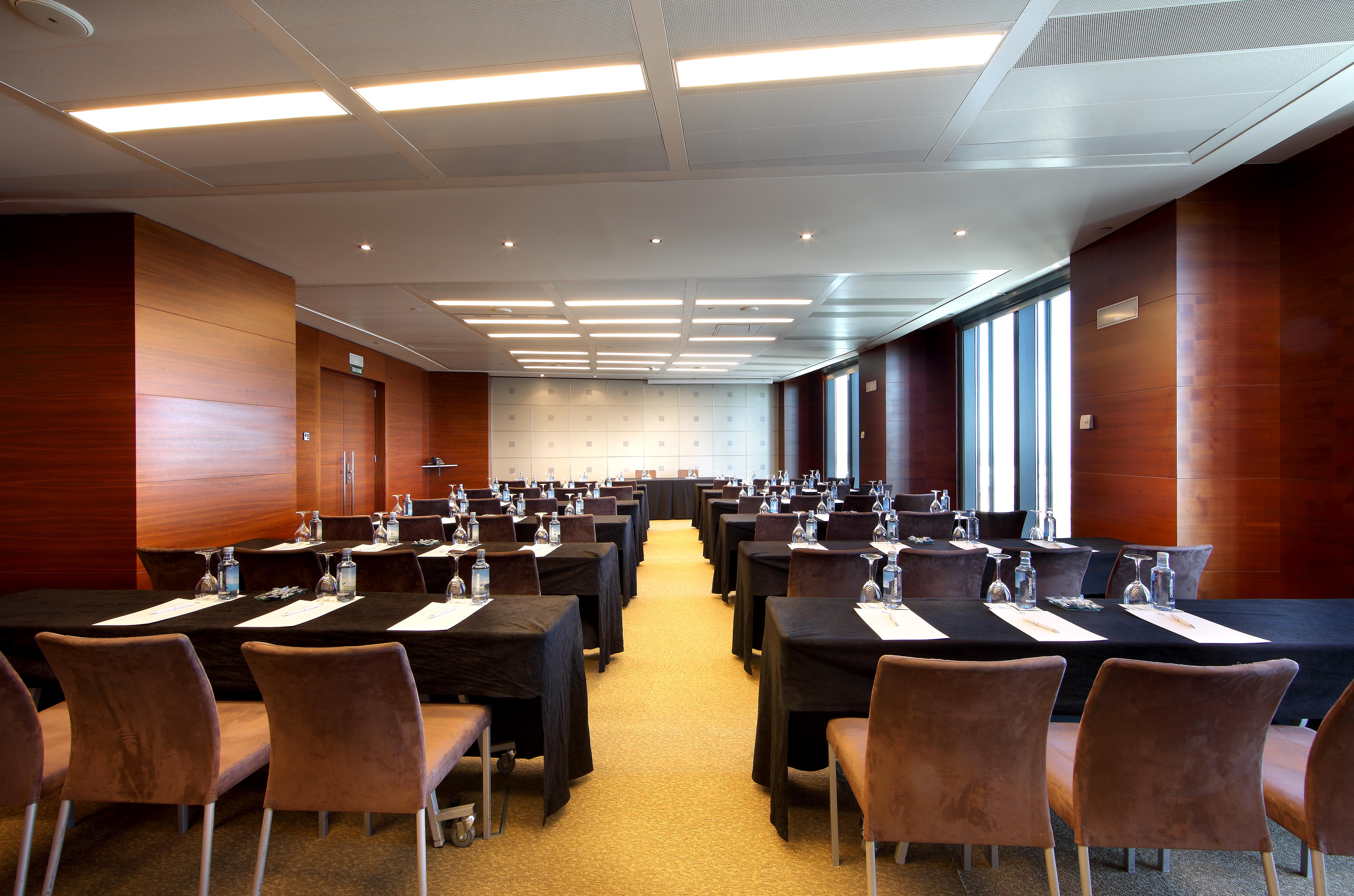 유로스타 마드리드 타워 외부 사진 The photo shows a modern conference room set up for a meeting or seminar. There are multiple tables arranged in rows, each covered with black tablecloths. Each table has a clean white sheet, and water bottles are placed at each setting. The room is w