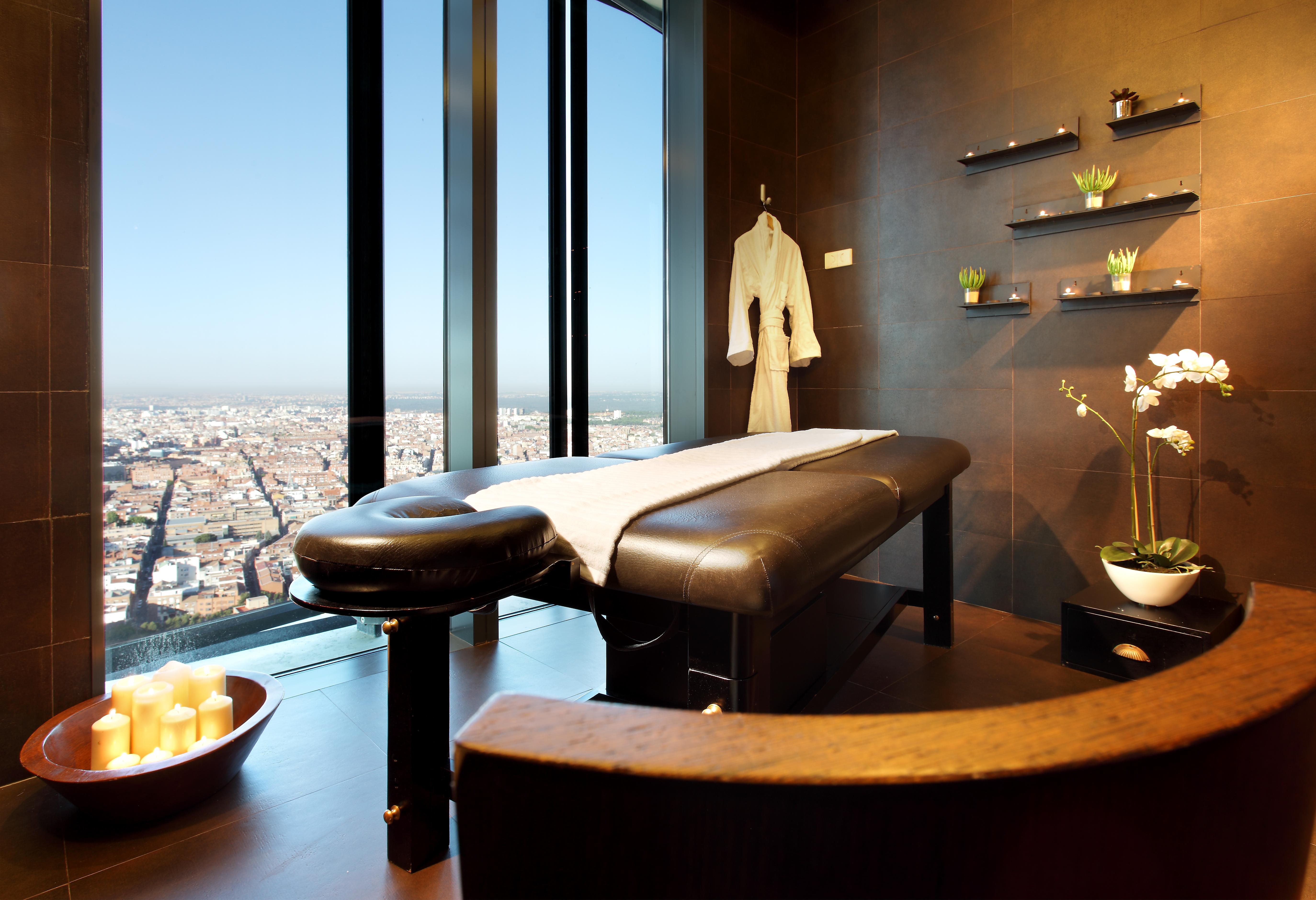 유로스타 마드리드 타워 외부 사진 The photo shows a modern spa treatment room with large windows offering a panoramic view of the city. In the center, there's a massage table covered with a white sheet. A plush bathrobe hangs on the wall, and there are decorative shelves with small p