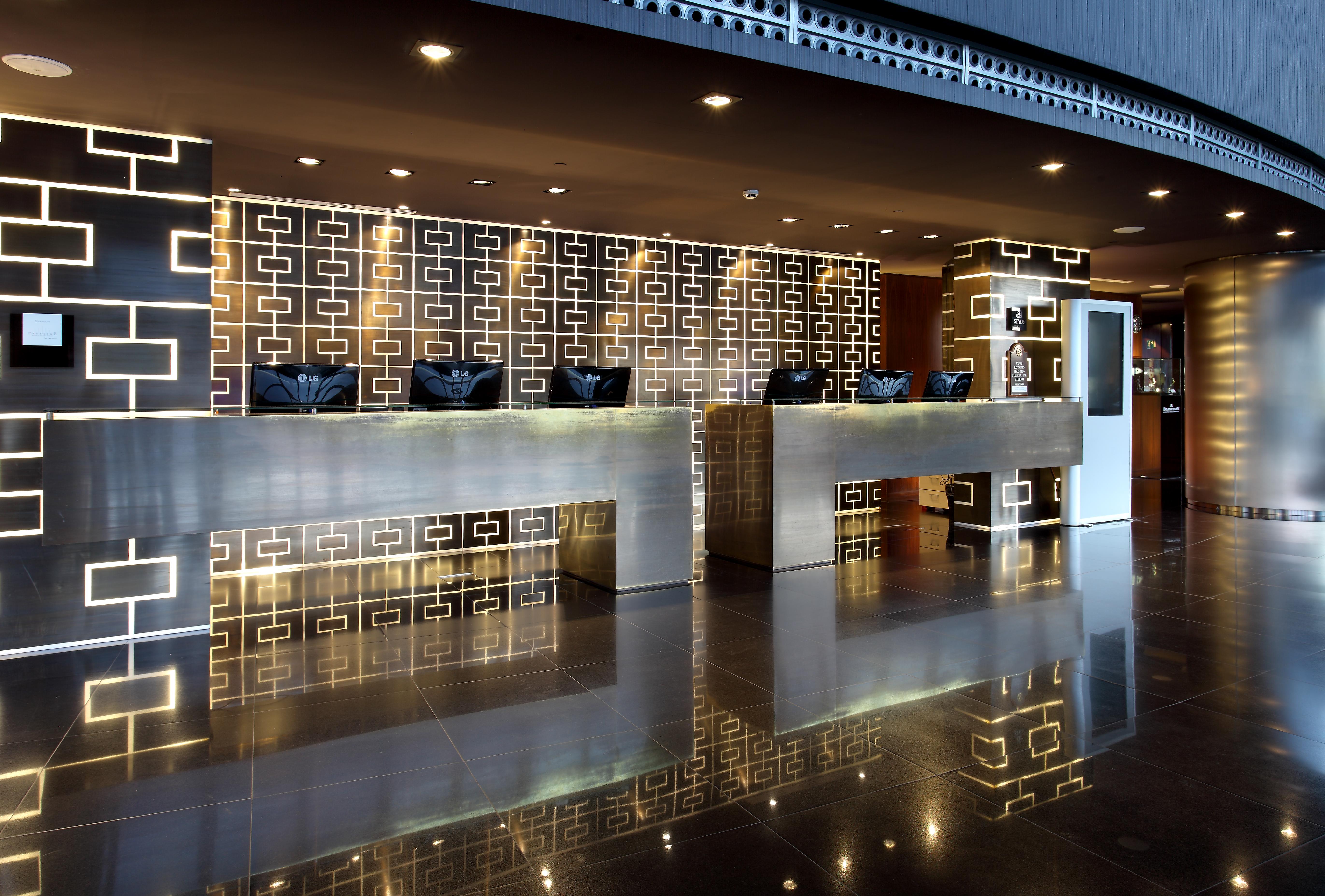 유로스타 마드리드 타워 외부 사진 The photo shows a modern hotel reception area. There are two prominent reception counters made of a sleek, shiny material. The background features a decorative wall with a geometric pattern, possibly in darker colors. The lighting is soft and stylish