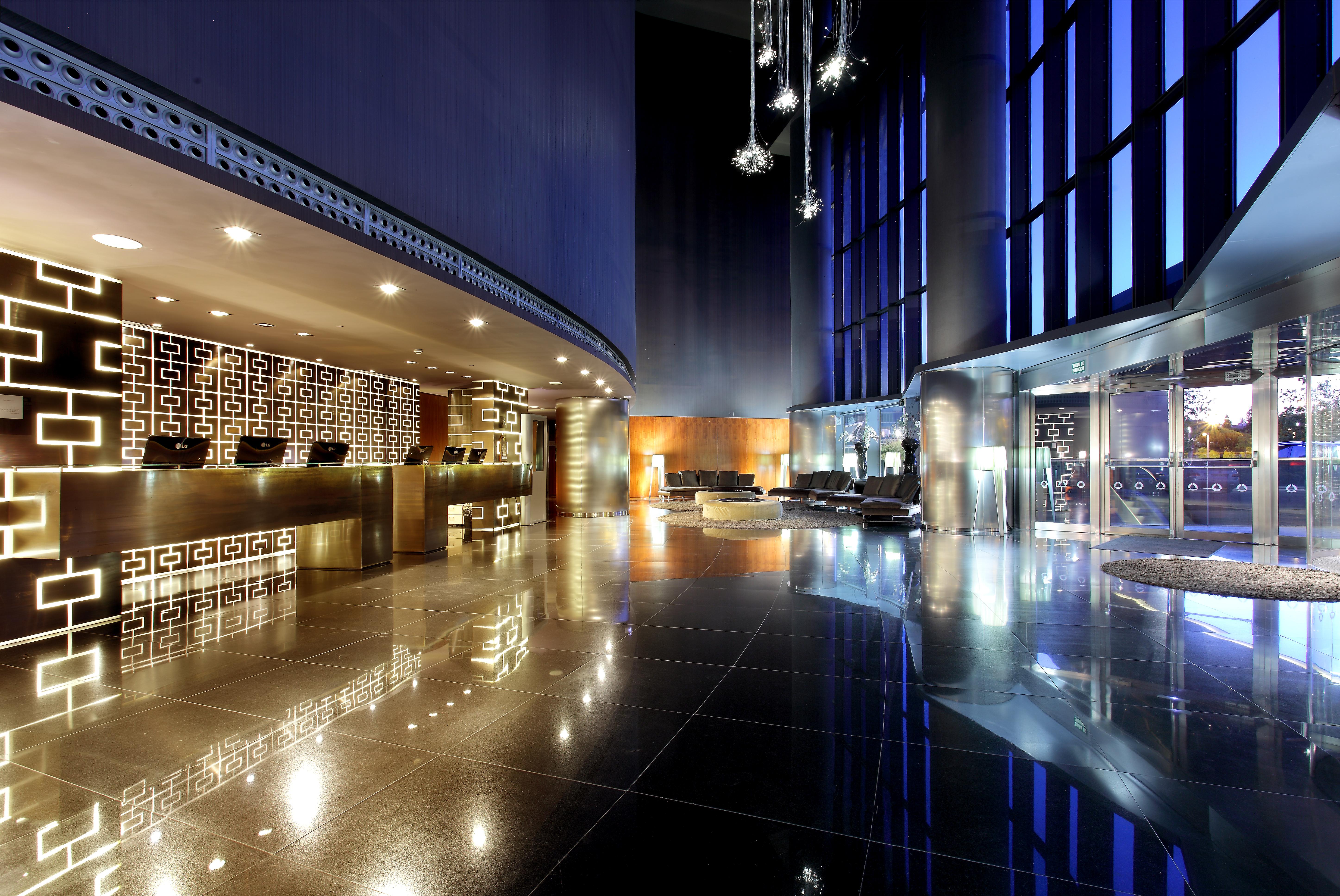 유로스타 마드리드 타워 외부 사진 The image depicts a modern hotel lobby featuring sleek, contemporary design elements. On one side, there is a reception desk with a striking geometric pattern, illuminated with soft lighting. The flooring is glossy and reflective, adding to the elega
