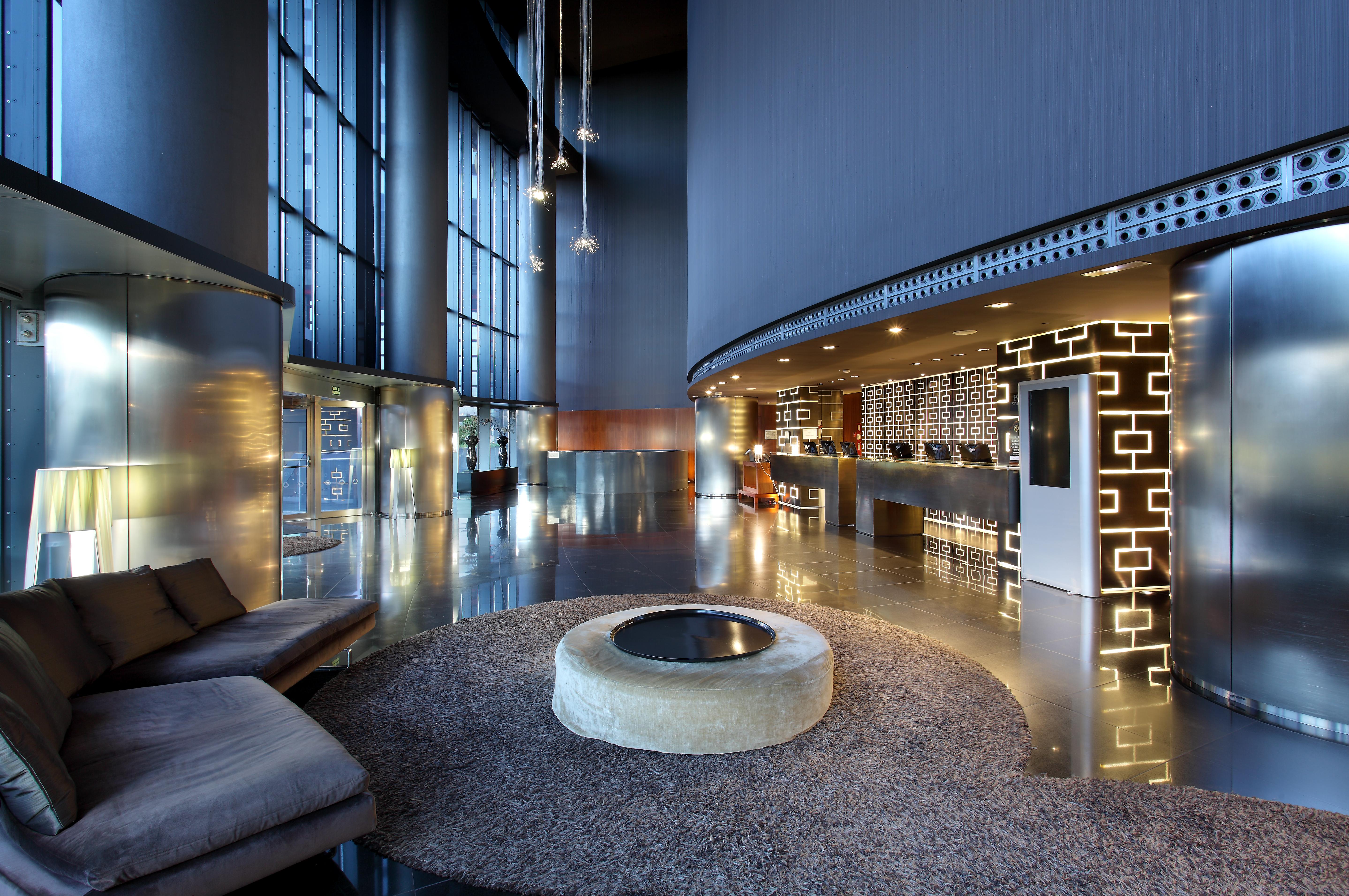 유로스타 마드리드 타워 외부 사진 The photo shows a modern hotel lobby with a sleek and sophisticated design. There is a large, open space featuring tall glass windows allowing natural light to flood in. The reception desk is stylishly designed, with a decorative wall behind it that 