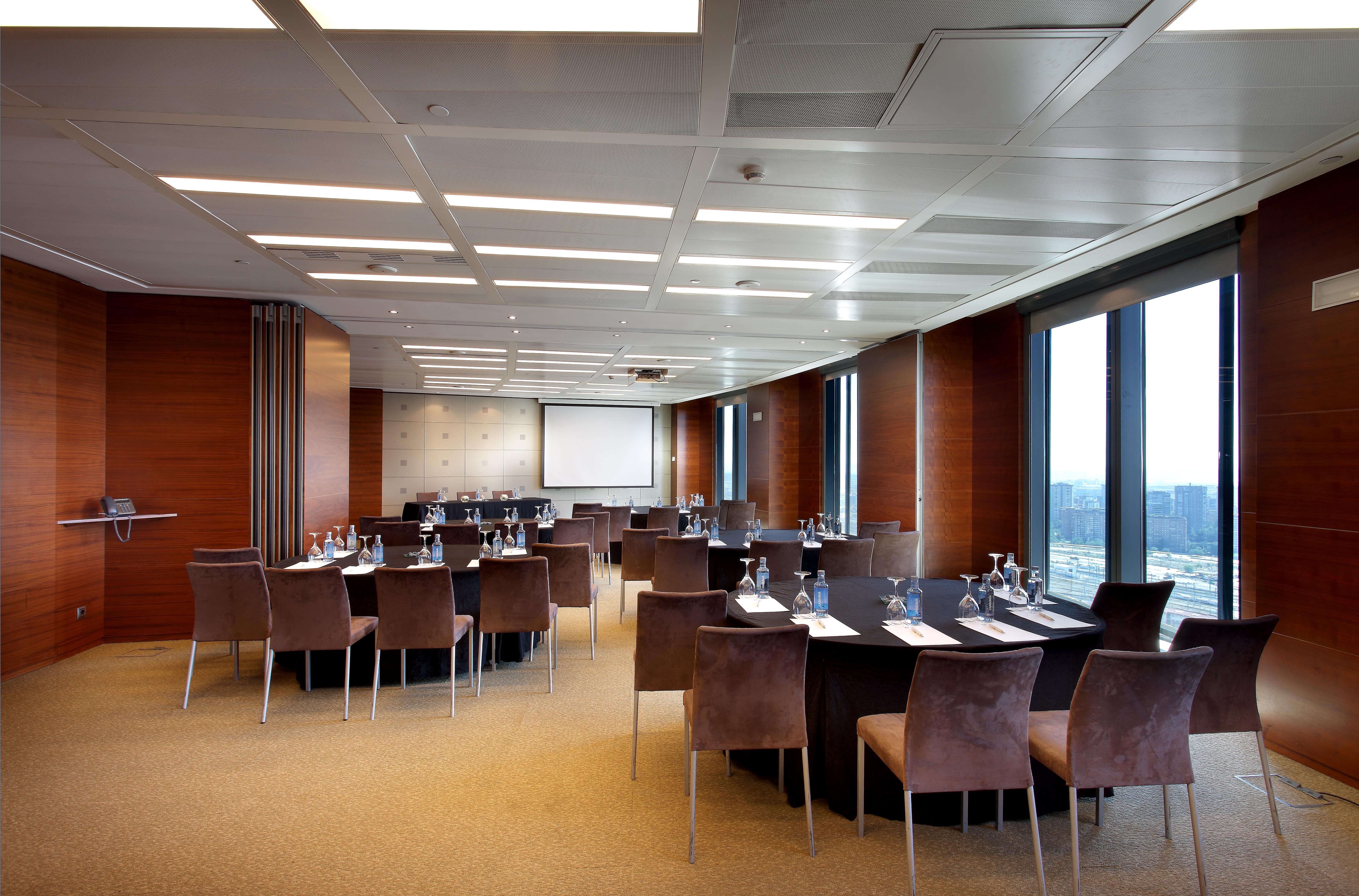 유로스타 마드리드 타워 외부 사진 The photo shows a modern conference room set up for a meeting or event. The room features a large, rectangular table surrounded by upholstered chairs. Each place at the table has a notepad and pen, and there are glasses of water provided. Large windo