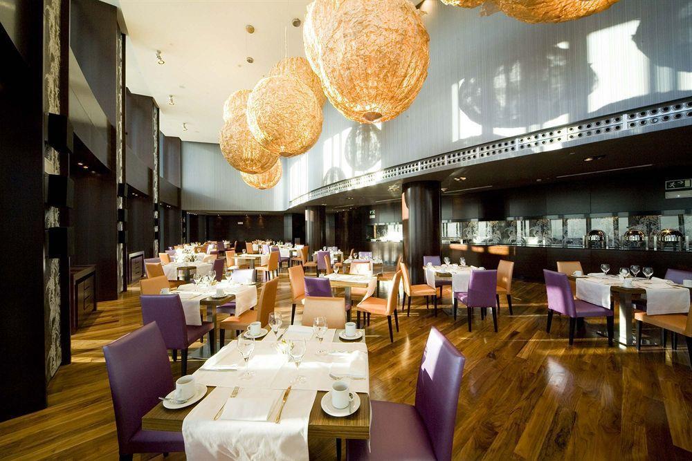 유로스타 마드리드 타워 레스토랑 사진 The photo shows a modern restaurant interior featuring elegant decor. The dining area includes tables set for guests with white tablecloths, plates, and cutlery. The seating consists of a mix of wooden chairs, some upholstered in purple fabric. The l
