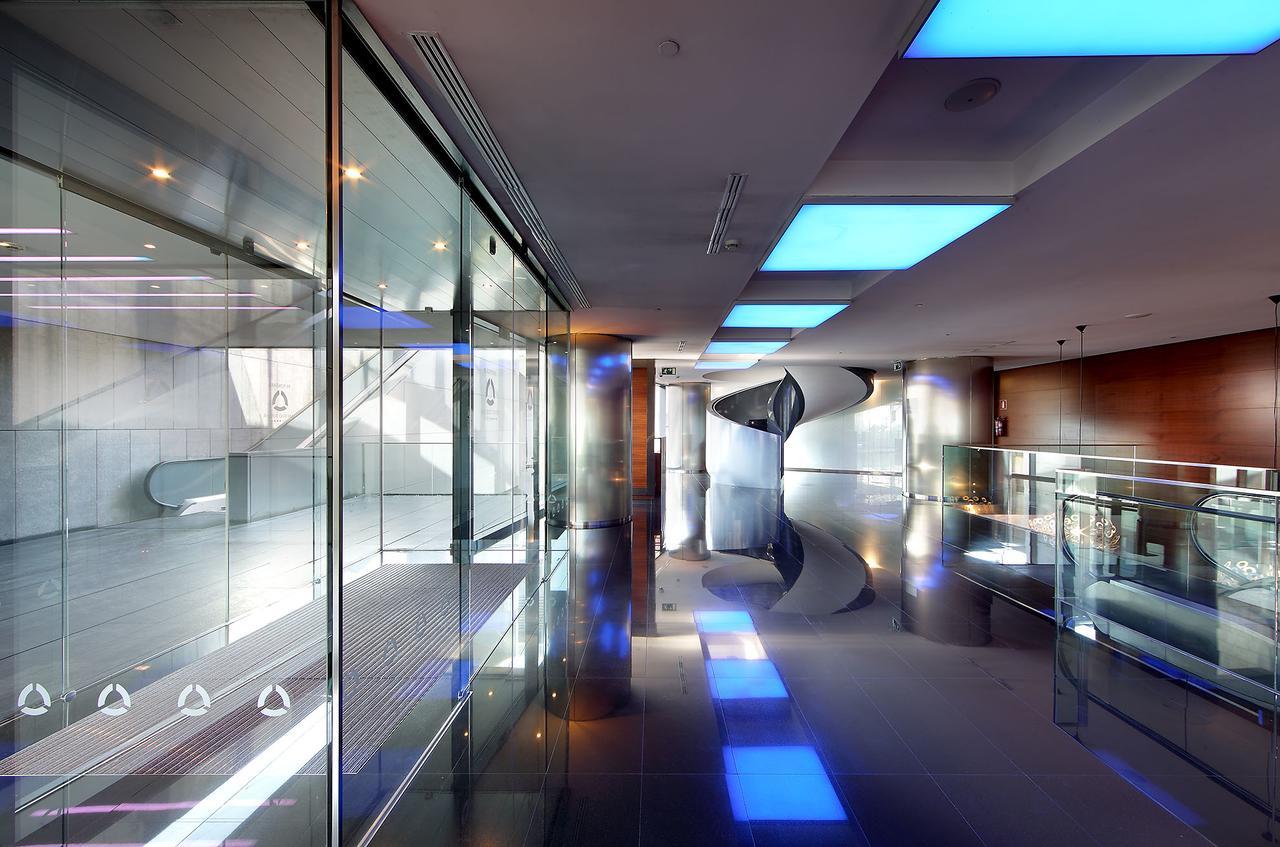 유로스타 마드리드 타워 외부 사진 The photo shows a modern, sleek interior space characterized by large glass walls and reflective surfaces. The ceiling features blue-lit sections, creating a contemporary atmosphere. The polished floor enhances the overall brightness of the area, and