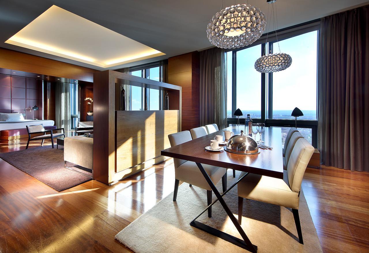 유로스타 마드리드 타워 외부 사진 The photo shows a modern dining area within a spacious interior. A sleek wooden dining table is set for a meal, featuring white dining chairs. Above the table, there are stylish chandelier lights illuminating the space. Large floor-to-ceiling windows