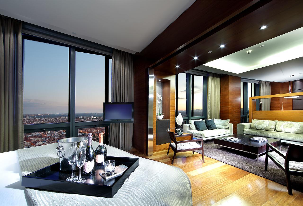 유로스타 마드리드 타워 외부 사진 The photo shows a luxurious hotel suite with modern decor. On one side, there is a bedroom area featuring a large bed, while on the other side, there is a spacious living area with a comfortable sofa and coffee table. The room is well-lit with stylis