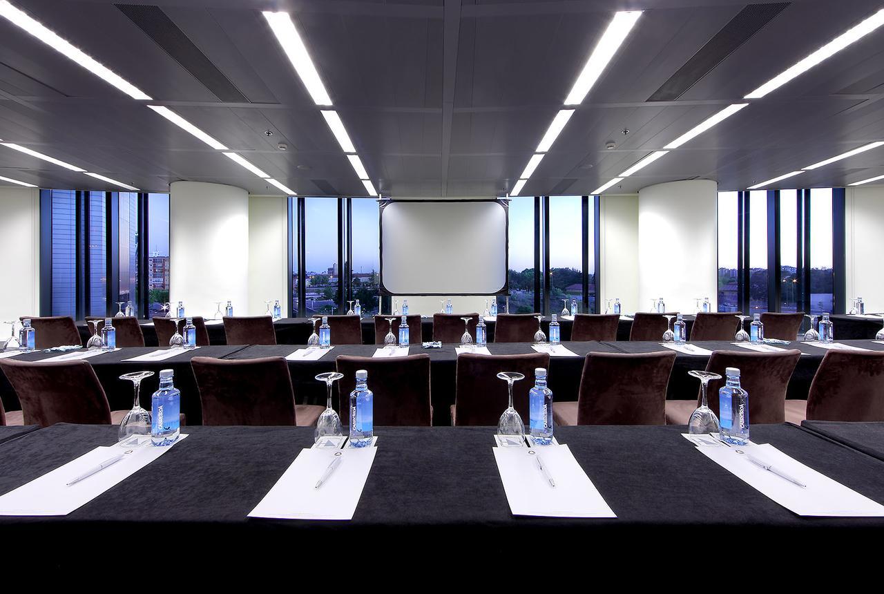 유로스타 마드리드 타워 외부 사진 The photo shows a modern conference room with large windows that provide a view of the cityscape outside. The room is arranged with several tables set up in a classroom or boardroom style. Each table has a black tablecloth and features a glass of wat