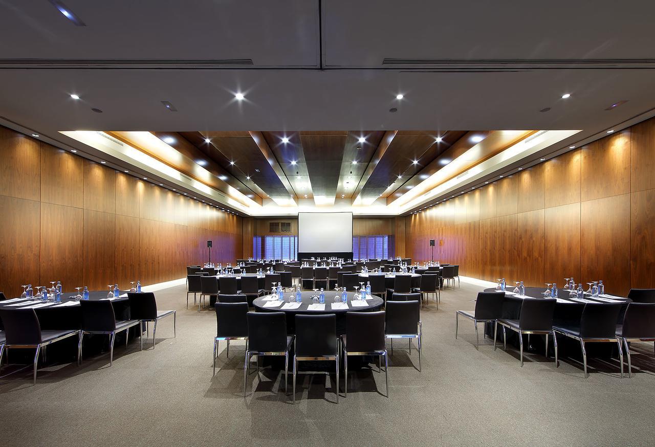 유로스타 마드리드 타워 외부 사진 The photo shows a spacious and modern conference room set up for a meeting or event. There are several round tables arranged in a semi-circle, each covered with a white tablecloth and equipped with water glasses, writing pads, and pens. The room feat