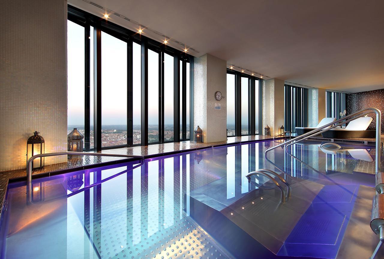 유로스타 마드리드 타워 외부 사진 The photo shows a luxurious indoor pool area with large floor-to-ceiling windows that provide a stunning view of the skyline. The pool features a shiny, reflective surface and has a modern design, with gentle curves in the structure. The lighting is 