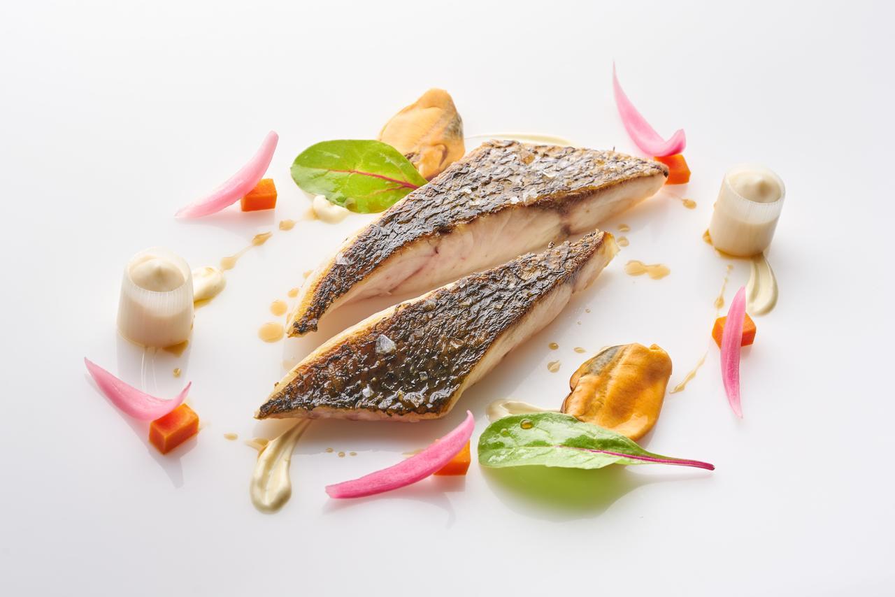 유로스타 마드리드 타워 외부 사진 The image depicts a beautifully plated dish featuring two pieces of fish fillet, likely a type of white fish, arranged artfully on a white plate. The fish has a crispy skin and is cooked to a tender texture. Surrounding the fish are various garnishes