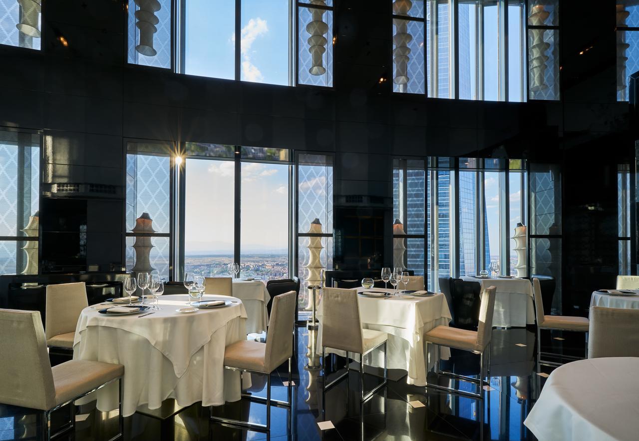 유로스타 마드리드 타워 외부 사진 The photo features an elegant restaurant interior with large windows that provide a panoramic view of the city skyline. The tables are elegantly set with white tablecloths and fine dining tableware, including glasses and plates. The decor appears mod