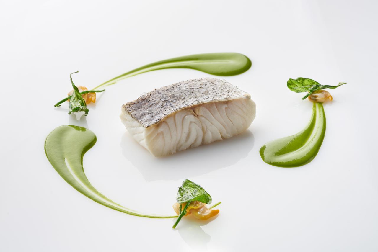 유로스타 마드리드 타워 외부 사진 The photo shows a beautifully presented dish featuring a piece of fish, likely a white fish, with a prominent skin side on top. It is surrounded by artistic garnishes, including vibrant green sauces and small sprigs of herbs. The composition is elega