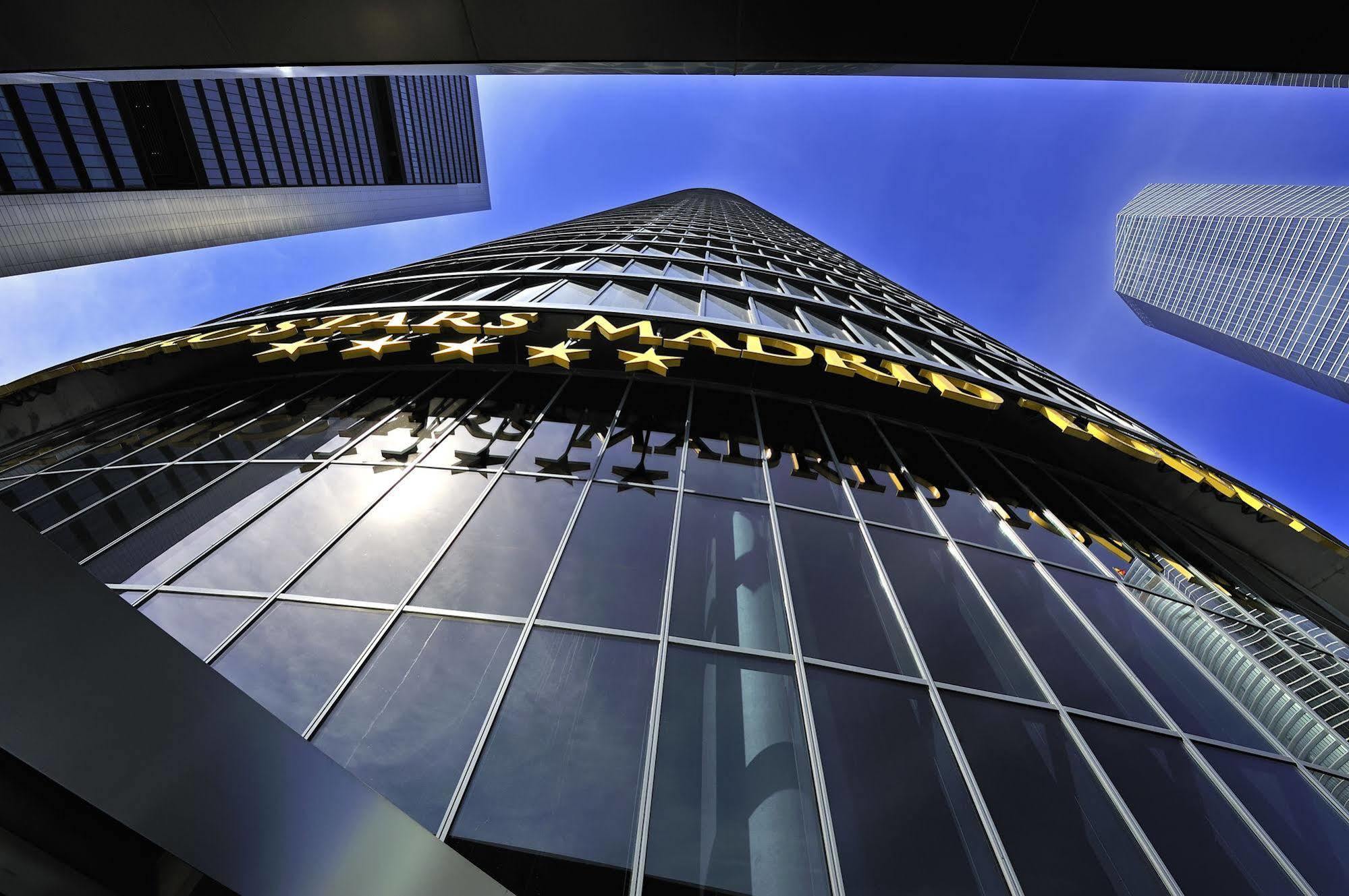 유로스타 마드리드 타워 외부 사진 The photo shows a tall modern building reaching towards the sky. The perspective is from the ground looking upward, highlighting the building's height and glass facade. The building features a large sign that reads "MADRID" in bright yellow letters, 
