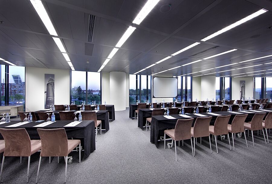 유로스타 마드리드 타워 외부 사진 The photo shows a modern conference room set up for a meeting or seminar. There are multiple rows of tables arranged in a classroom style, each covered with black tablecloths. On the tables, there are notepads and water bottles, suggesting that atten