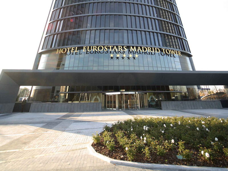 유로스타 마드리드 타워 외부 사진 The photo shows the exterior of the Eurostars Madrid Tower Hotel, which is a modern, high-rise building. The hotel features a sleek glass facade and has "HOTEL EUROSTARS MADRID TOWER" prominently displayed in gold lettering above the entrance. The ar